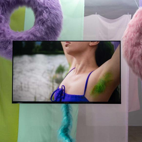 haarige, pastellfarbne Schwimmringe, Bildschirm auf dem eine Person zu sehen ist, die ihre Arme in die Luft streckt, giftgrüne Achselhaare zeigt und ein lila Bikini-Oberteil trägt