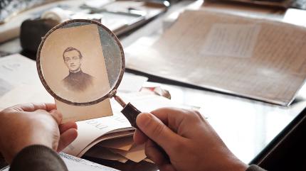 H&auml;nde halten eine Lupe &uuml;ber ein altes, vergilbtes Foto auf einem Schreibtisch mit weiteren Papieren und Dokumenten.