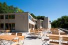 Flacher moderner Geb&auml;udekomplex mit gro&szlig;en Fenstern und mehreren Holz-Gartentischen mit St&uuml;hlen auf einer Terrasse unter blauem Himmel