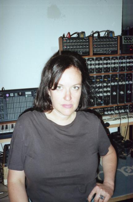 Eine weiblich gelesene Person mit einer Wand mit Synthesizern im Rücken.