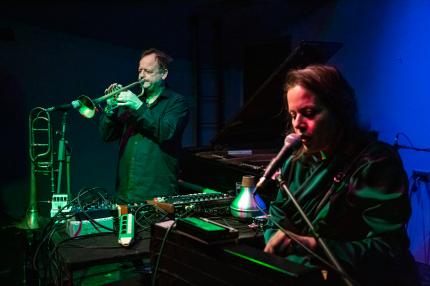 Eine männlich gelesene Person spielt Trompete und steht links, rechts davon eine weiblich gelesene Person, die in ein Mikrofon Singt und Synthesizer spielt. Zwischen Ihnen eine Menge Instrumente, wie Synthesizer, Posaune, Melodica und Effektgeräte.