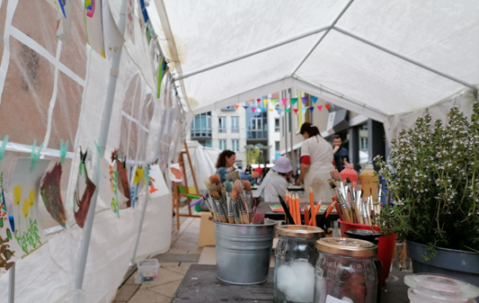 Das Bild zeigt ein Zelt, in dem auf einem Tisch Farbdosen mit Pinseln darin zu sehen sind.