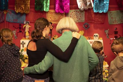 Zwei Menschen stehen Arm im Arm in einem Ausstellungsraum zum Thema "Mexikanisches Totenfest“ mit bunten Girlanden und Totenköpfen
