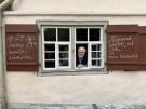 Ein Mann schaut aus dem Sprossenfenster eines Hauses. Die Fensterl&auml;den sind beschrieben.