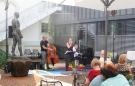 Auf dem Bild ist eine dreiköpfige Band zu sehen, die mit Kontrabass und Klavier eine Sängerin begleitet. Sie stehen in dem Innenhof des Edwin Scharff Museums neben einer Skultpur, im Vordergrund ist Publikum zu sehen.