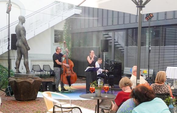 Auf dem Bild ist eine dreik&ouml;pfige Band zu sehen, die mit Kontrabass und Klavier eine S&auml;ngerin begleitet. Sie stehen in dem Innenhof des Edwin Scharff Museums neben einer Skultpur, im Vordergrund ist Publikum zu sehen.