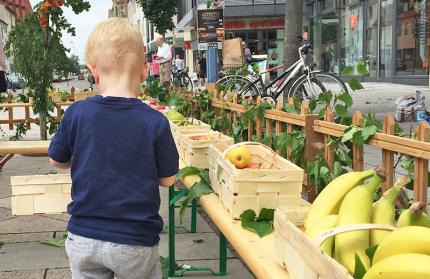 Ein Kind steht mit einem Korb neben Obstkisten mit Banen und &Auml;pfeln.