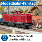 Rote Diesellokomotive der Baureihe 211 im Ma&szlig;stab H0 steht vor einem kleinen Bahnhofsgeb&auml;ude auf einer Modellbahnanlage. Oben im Bild ein blauer Banner mit der Aufschrift &bdquo;Modellbahn-Fahrtag&ldquo;, unten das Logo und der Name &bdquo;Modellbahnfreunde Ulm/Neu-Ulm e.V.&ldquo;.