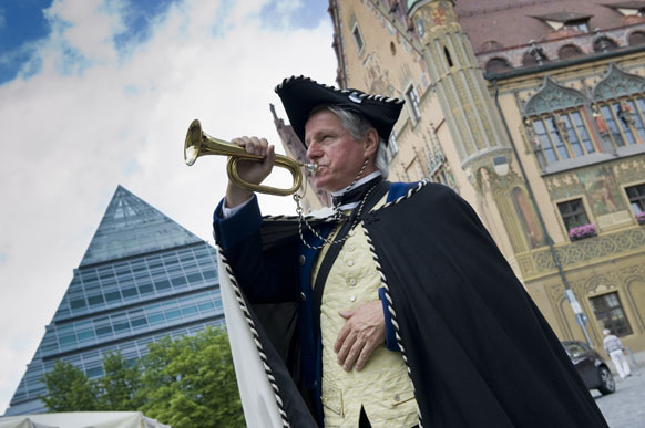 Gästeführer in historischem Gewand mit historischem Blasinstrument vor Ulmer Glaspyramide und historischem Rathaus