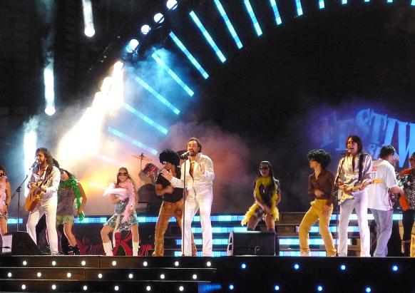 Abbildung einer Musikalische Szene der Show "Massachusetts - Bee Gees Musical" mit der Band im Vordergrund der Bühne, stehend an Mikrofonen und Tänzern im Hintergrund