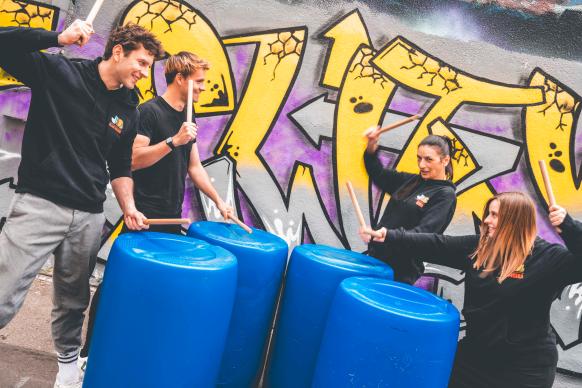 Das Ensemble der JUB beim Trommeln auf Plastikfässern vor einer Grafitti-Wand