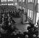Schwarz-wei&szlig;e Aufnahme einer gro&szlig;en Menschenmenge in einem Saal, die einem Redner an einem Podium mit Mikrofonen zuh&ouml;rt