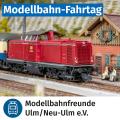 Modellbahn-Fahrtag: Eine rote Diesellok (Baureihe 211) fährt auf einer detailreichen Modellbahnanlage an einem Gebäude im Alpenstil vorbei. Zwei Modellfiguren sitzen auf einer Bank vor dem Haus. Oben steht in einem blauen Balken „Modellbahn-Fahrtag“. Unten das Logo der Modellbahnfreunde Ulm/Neu-Ulm e.V.