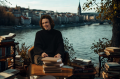 Ein l&auml;chelnder Mann vor einigen Stapeln B&uuml;cher. Im Hintergrund sieht man einen Fluss sowie eine Stadt.
